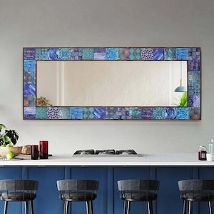 May include: A large rectangular mirror with a blue and brown mosaic tile frame. The mirror is hanging above a kitchen counter with a blue cabinet and three black bar stools.