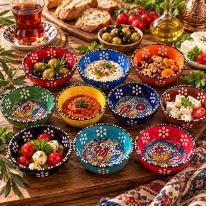Puede incluir: Una colección de diez cuencos de cerámica pequeños y redondos con vibrantes patrones florales multicolores y motivos de pétalos blancos en los bordes. Los cuencos están dispuestos sobre una superficie de madera, algunos con aceitunas, salsas, frutos secos, queso y tomates.