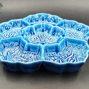 May include: A blue ceramic serving dish with a floral design. The dish is divided into seven sections, each with a detailed white pattern. The edges are scalloped, and the overall shape is reminiscent of a flower. The dish is approximately 12 inches in diameter.