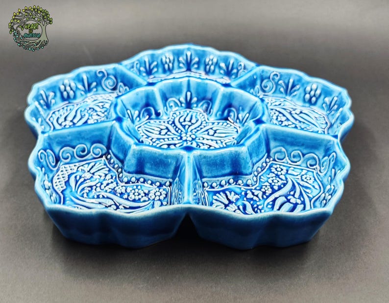 May include: A blue ceramic serving dish with a floral design. The dish is divided into seven sections, each with a detailed white pattern. The edges are scalloped, and the overall shape is reminiscent of a flower. The dish is approximately 12 inches in diameter.