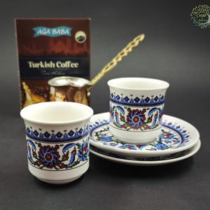 May include: Two white ceramic cups and two matching saucers decorated with blue, red, and green floral patterns. A metallic coffee pot with a long handle sits behind the cups next to a brown box labeled Ağa Baba Turkish Coffee.