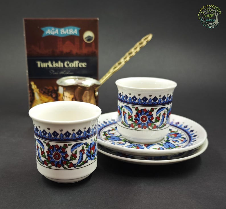 May include: Two white ceramic cups and two matching saucers decorated with blue, red, and green floral patterns. A metallic coffee pot with a long handle sits behind the cups next to a brown box labeled Ağa Baba Turkish Coffee.