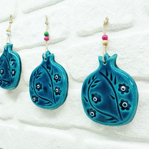 Peut inclure: Trois ornements en céramique de grenade bleu sarcelle avec des motifs floraux blancs et noirs et des détails de mauvais œil suspendus à une ficelle avec des perles colorées.