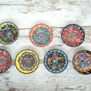 Puede incluir: Una colecci&oacute;n de siete cuencos peque&ntilde;os y redondos de cer&aacute;mica con intrincados patrones florales y geom&eacute;tricos coloridos. Cada cuenco presenta un dise&ntilde;o &uacute;nico con un borde negro, amarillo, naranja, verde, rojo o azul. Los cuencos est&aacute;n dispuestos sobre una superficie de madera blanca envejecida.