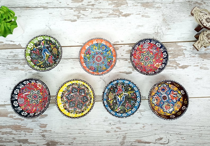 Puede incluir: Una colecci&oacute;n de siete cuencos peque&ntilde;os y redondos de cer&aacute;mica con intrincados patrones florales y geom&eacute;tricos coloridos. Cada cuenco presenta un dise&ntilde;o &uacute;nico con un borde negro, amarillo, naranja, verde, rojo o azul. Los cuencos est&aacute;n dispuestos sobre una superficie de madera blanca envejecida.