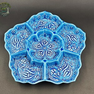 May include: A turquoise ceramic serving dish with a floral design. The dish is divided into six sections, with a central flower-shaped compartment. The intricate white floral patterns contrast with the blue background. The dish has a scalloped edge.