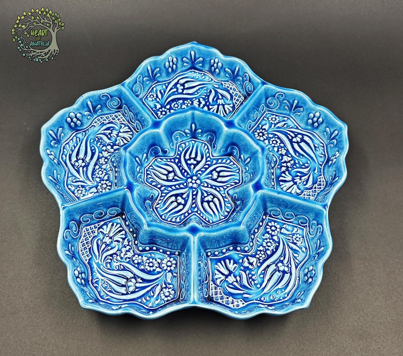 May include: A turquoise ceramic serving dish with a floral design. The dish is divided into six sections, with a central flower-shaped compartment. The intricate white floral patterns contrast with the blue background. The dish has a scalloped edge.
