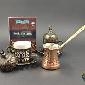 May include: A copper Turkish coffee pot with a long handle, two ornate metal cup holders with lids, two white ceramic inserts, and a box labeled AGA BABA Turkish Coffee. The metal components feature intricate filigree patterns and crescent moon finials.