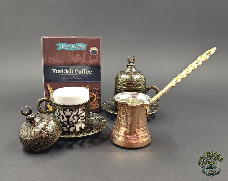 May include: A copper Turkish coffee pot with a long handle, two ornate metal cup holders with lids, two white ceramic inserts, and a box labeled AGA BABA Turkish Coffee. The metal components feature intricate filigree patterns and crescent moon finials.