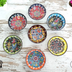 Puede incluir: Una colecci&oacute;n de siete cuencos peque&ntilde;os y redondos de cer&aacute;mica con intrincados y coloridos dise&ntilde;os florales. Cada cuenco presenta un dise&ntilde;o &uacute;nico con una variedad de colores, incluyendo rojo, azul, amarillo y verde. Los cuencos est&aacute;n dispuestos sobre una superficie de madera clara.