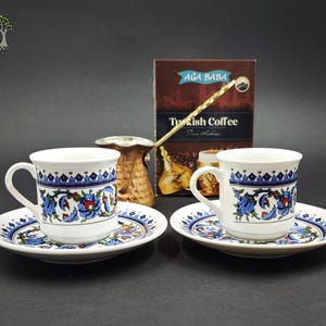 May include: Two white ceramic coffee cups with matching saucers, each featuring a blue, red, and green floral pattern. A copper-colored metal coffee pot sits behind the cups, next to a brown box labeled AGA BABA Turkish Coffee.