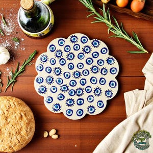 Può includere: Sottopentola in ceramica bianca con un design di occhio turco blu e bianco. Il sottopentola ha la forma di un fiore con bordi arrotondati.