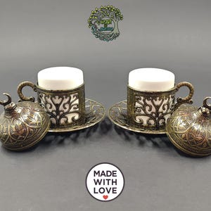 May include: Two white ceramic cups set in ornate, antique brass-colored metal holders with matching saucers and domed lids featuring crescent moon finials. The metalwork displays intricate filigree patterns. Text includes Heart of Anatolia and Made With Love.