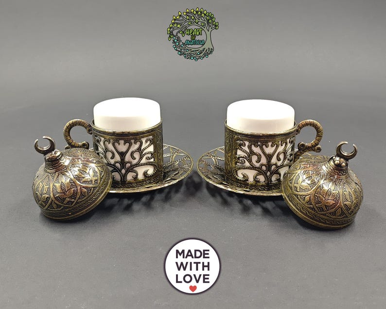 May include: Two white ceramic cups set in ornate, antique brass-colored metal holders with matching saucers and domed lids featuring crescent moon finials. The metalwork displays intricate filigree patterns. Text includes Heart of Anatolia and Made With Love.