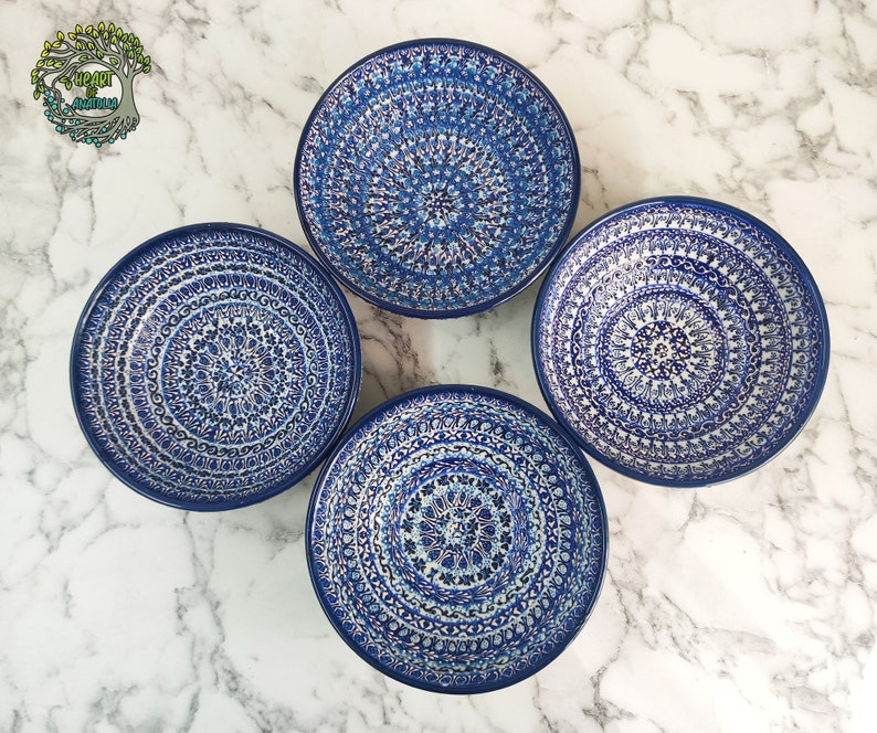 May include: Four blue and white ceramic plates with intricate, symmetrical patterns. The plates have a dark blue rim and a detailed, floral-like design in shades of blue and white. The plates are arranged on a white and gray marble surface.
