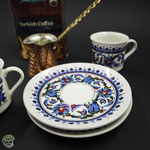 May include: A white ceramic saucer and matching cup featuring a blue, red, and green floral pattern. A copper-colored metal Turkish coffee pot with a long handle sits behind the saucer, next to a box labeled Turkish Coffee in white text.