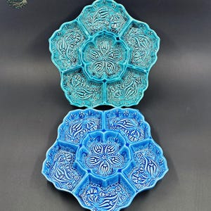 May include: Two ceramic serving dishes in a flower shape, one turquoise and one blue. Each dish has six compartments around a central well, with floral and leaf patterns in white. The dishes are on a dark gray background.