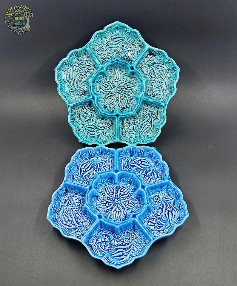 May include: Two ceramic serving dishes in a flower shape, one turquoise and one blue. Each dish has six compartments around a central well, with floral and leaf patterns in white. The dishes are on a dark gray background.