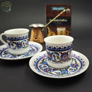 May include: Two white ceramic coffee cups and matching saucers decorated with blue, red, and green floral patterns. A copper-colored metal coffee pot with a long handle sits behind the cups, alongside a brown box labeled Turkish Coffee.