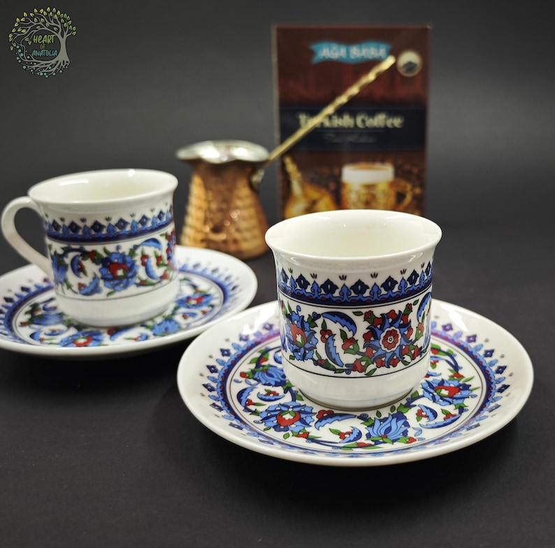 May include: Two white ceramic coffee cups and matching saucers decorated with blue, red, and green floral patterns. A copper-colored metal coffee pot with a long handle sits behind the cups, alongside a brown box labeled Turkish Coffee.