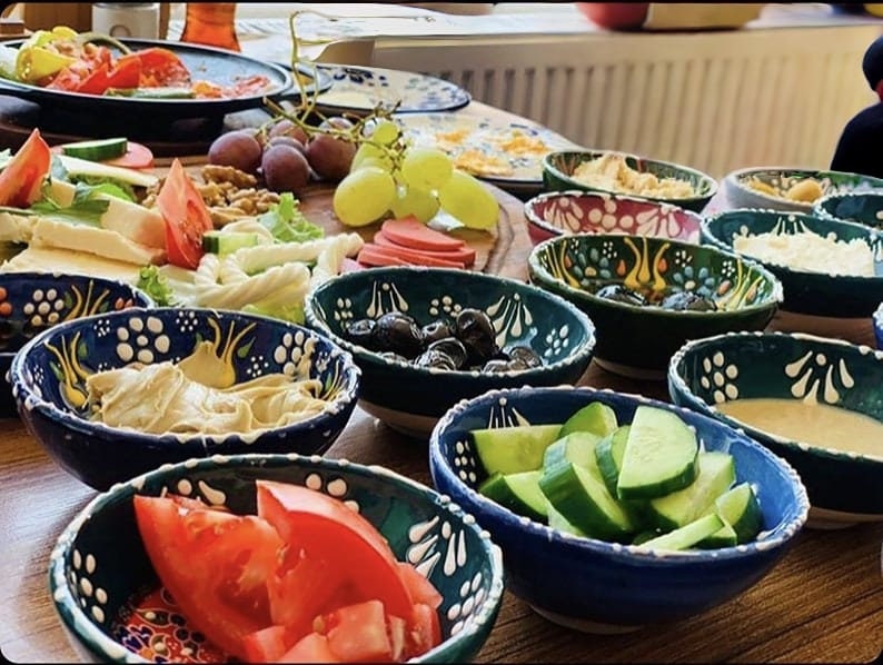 May include: A table laden with a variety of breakfast foods. Several small, decorative bowls filled with olives, sliced cucumbers, tomatoes, and dips. Other plates hold cheese, fruit, and nuts. The bowls have a blue and white floral design.