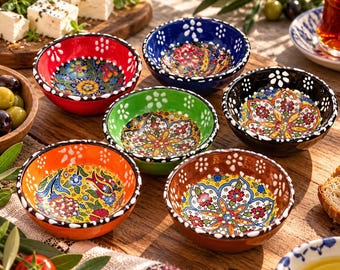 Może przedstawiać: Zestaw sześciu małych ceramicznych misek z żywymi, ręcznie malowanymi wzorami kwiatowymi w kolorach czerwonym, niebieskim, zielonym, czarnym, pomarańczowym i brązowym. Każda miska ma biały kropkowany brzeg i złożone, kolorowe motywy roślinne.