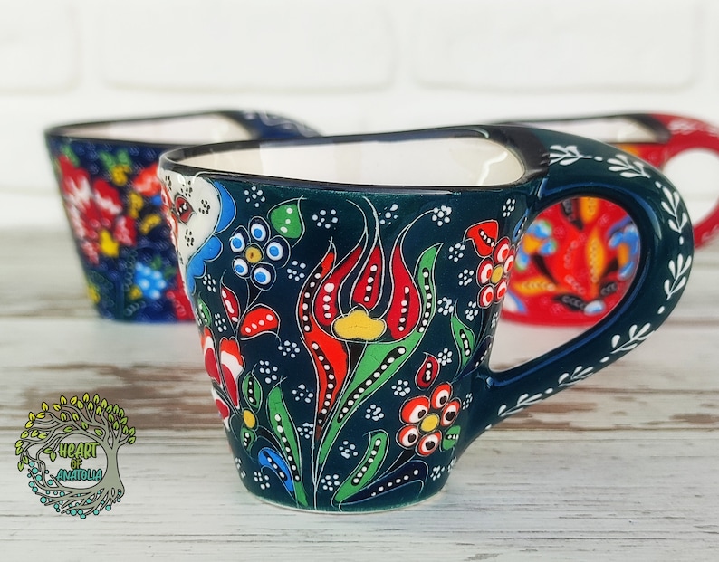May include: A set of three Turkish coffee cups with floral designs. The cups are made of ceramic and have a dark blue background with colorful flowers and leaves. The cups have a handle on the side and are perfect for serving Turkish coffee.