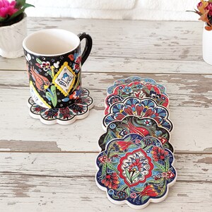 Puede incluir: Una taza de cer&aacute;mica negra con un dise&ntilde;o floral se encuentra sobre un posavasos a juego. Varios posavasos adicionales con un patr&oacute;n floral similar est&aacute;n apilados cerca. Los posavasos y la taza presentan patrones florales rojos, azules y verdes.