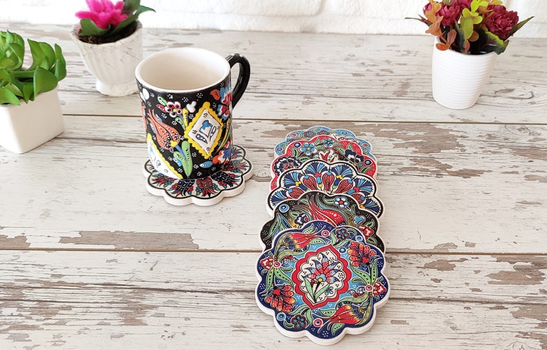 Puede incluir: Una taza de cer&aacute;mica negra con un dise&ntilde;o floral se encuentra sobre un posavasos a juego. Varios posavasos adicionales con un patr&oacute;n floral similar est&aacute;n apilados cerca. Los posavasos y la taza presentan patrones florales rojos, azules y verdes.