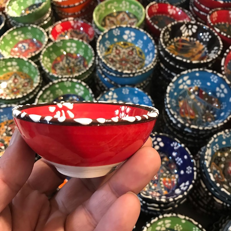 May include: A small, red ceramic bowl with a white floral pattern around the rim. The bowl is held in a hand, with many other colorful bowls stacked in the background. The bowls have various colors and patterns, including blue, green, and orange.