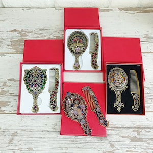 May include: Ornate vanity mirror and comb sets in red gift boxes. The mirrors and combs have intricate designs with colorful jewel accents. The sets feature peacock and floral motifs, with a vintage aesthetic. The sets are displayed on a light wood-grain surface.