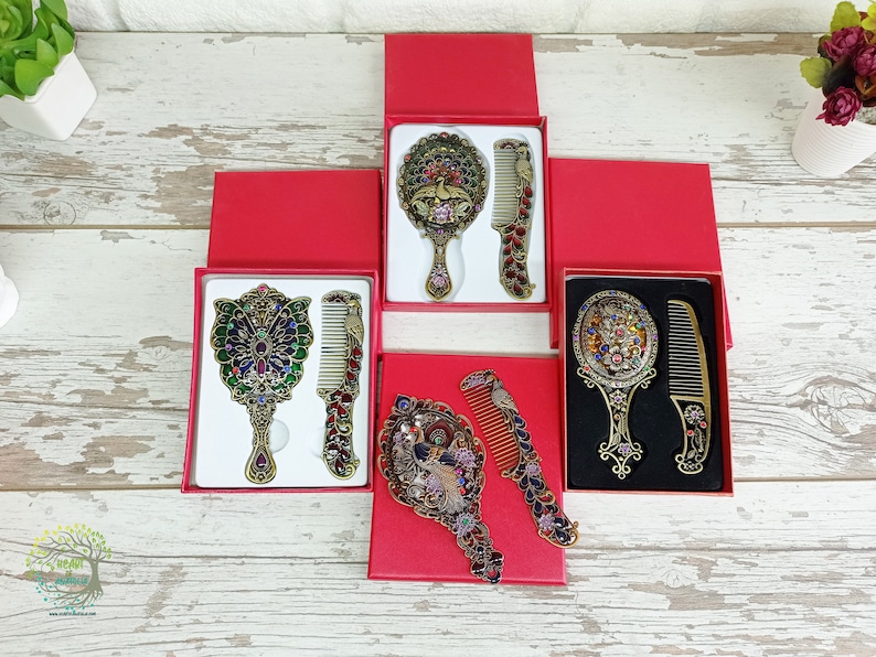 May include: Ornate vanity mirror and comb sets in red gift boxes. The mirrors and combs have intricate designs with colorful jewel accents. The sets feature peacock and floral motifs, with a vintage aesthetic. The sets are displayed on a light wood-grain surface.