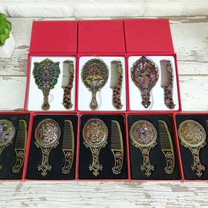 May include: Ornate antique-style mirror and comb sets in red boxes. The sets feature detailed metalwork with colorful jewel accents. Each set includes a hand mirror and a matching comb, perfect for vanity or travel.