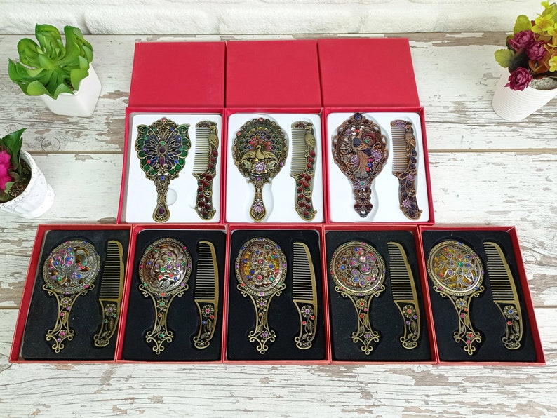 May include: Ornate antique-style mirror and comb sets in red boxes. The sets feature detailed metalwork with colorful jewel accents. Each set includes a hand mirror and a matching comb, perfect for vanity or travel.