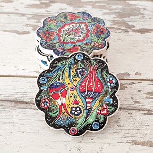Puede incluir: Una pila de posavasos decorativos de cer&aacute;mica con borde festoneado. Cada posavasos presenta un vibrante dise&ntilde;o floral en rojo, azul, amarillo y verde sobre un fondo blanco y negro. Los posavasos est&aacute;n apilados sobre una superficie de madera blanca envejecida.