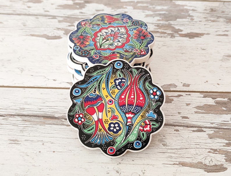 Puede incluir: Una pila de posavasos decorativos de cer&aacute;mica con borde festoneado. Cada posavasos presenta un vibrante dise&ntilde;o floral en rojo, azul, amarillo y verde sobre un fondo blanco y negro. Los posavasos est&aacute;n apilados sobre una superficie de madera blanca envejecida.