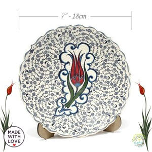 May include: A white ceramic decorative plate with a blue and red floral design. The plate is 7 inches in diameter and features a stylized tulip with intricate details. The plate is on a wooden stand.