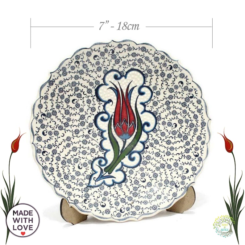 May include: A white ceramic decorative plate with a blue and red floral design. The plate is 7 inches in diameter and features a stylized tulip with intricate details. The plate is on a wooden stand.