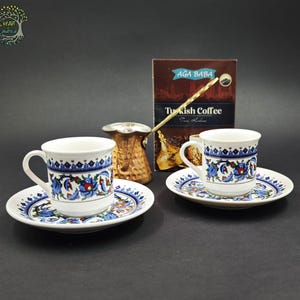 May include: Two white ceramic coffee cups with matching saucers, each featuring a blue and red floral pattern. A copper-colored metal coffee pot with a long handle sits between the cups, and a box labeled Aga Baba Turkish Coffee is in the back.