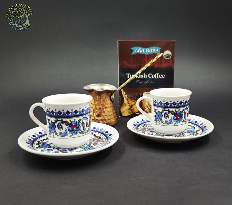 May include: Two white ceramic coffee cups with matching saucers, each featuring a blue and red floral pattern. A copper-colored metal coffee pot with a long handle sits between the cups, and a box labeled Aga Baba Turkish Coffee is in the back.