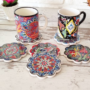 Puede incluir: Un juego de posavasos de cer&aacute;mica y dos tazas con dise&ntilde;os florales y paisley coloridos e intrincados. Los posavasos tienen un borde festoneado y est&aacute;n colocados sobre una superficie de madera clara. Las tazas tienen bordes negros y blancos.