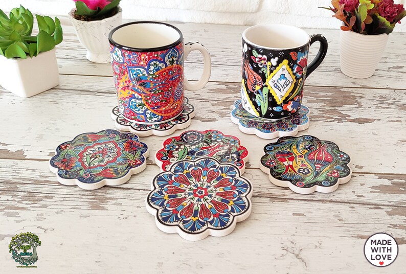 Puede incluir: Un juego de posavasos de cer&aacute;mica y dos tazas con dise&ntilde;os florales y paisley coloridos e intrincados. Los posavasos tienen un borde festoneado y est&aacute;n colocados sobre una superficie de madera clara. Las tazas tienen bordes negros y blancos.
