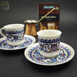 May include: Two white ceramic coffee cups and matching saucers decorated with blue, red, and green floral patterns. A copper-colored metal coffee pot with a long handle sits behind the cups, alongside a brown box labeled Turkish Coffee.
