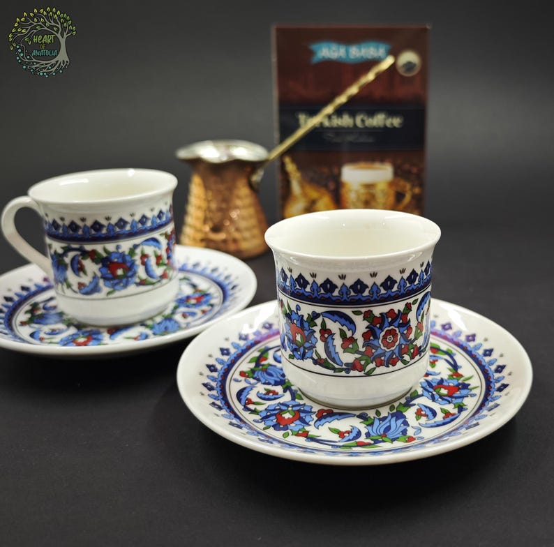 May include: Two white ceramic coffee cups and matching saucers decorated with blue, red, and green floral patterns. A copper-colored metal coffee pot with a long handle sits behind the cups, alongside a brown box labeled Turkish Coffee.