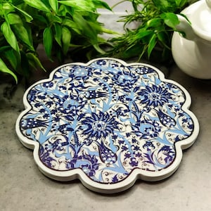 Puede incluir: Un posavasos de cerámica con borde festoneado, con un detallado estampado floral en tonos azules y blancos. El posavasos tiene un borde blanco y está diseñado para proteger las superficies del calor y la humedad. Decorativo y funcional.
