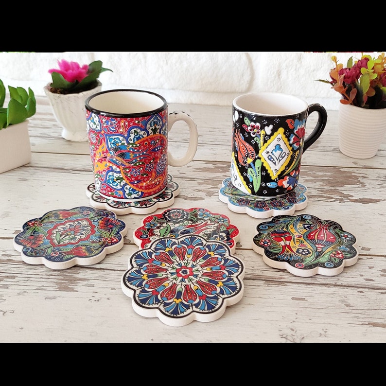 Puede incluir: Dos tazas de cer&aacute;mica y seis posavasos a juego con intrincados y coloridos dise&ntilde;os florales. Las tazas tienen asas y bordes blancos, mientras que los posavasos tienen forma de flor. La paleta de colores incluye rojo, azul y verde.