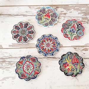 Puede incluir: Seis posavasos decorativos de cer&aacute;mica con dise&ntilde;os florales en rojo, azul y verde. Cada posavasos tiene un borde festoneado y un patr&oacute;n &uacute;nico, incluyendo flores estilizadas y formas geom&eacute;tricas. Los posavasos est&aacute;n colocados sobre una superficie de madera blanca envejecida.