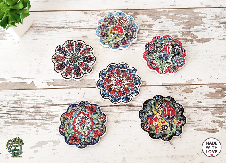 Puede incluir: Seis posavasos decorativos de cer&aacute;mica con dise&ntilde;os florales en rojo, azul y verde. Cada posavasos tiene un borde festoneado y un patr&oacute;n &uacute;nico, incluyendo flores estilizadas y formas geom&eacute;tricas. Los posavasos est&aacute;n colocados sobre una superficie de madera blanca envejecida.