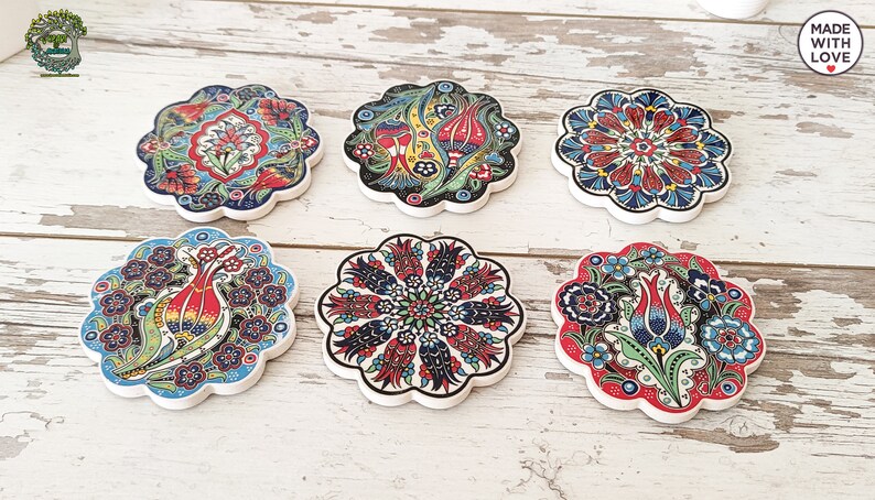 Puede incluir: Seis posavasos decorativos de cer&aacute;mica con dise&ntilde;os florales en rojo, azul, verde y amarillo. Cada posavasos tiene un borde festoneado y un dise&ntilde;o &uacute;nico. Est&aacute;n dispuestos sobre una superficie de madera blanca envejecida. La frase "MADE WITH LOVE" es visible.