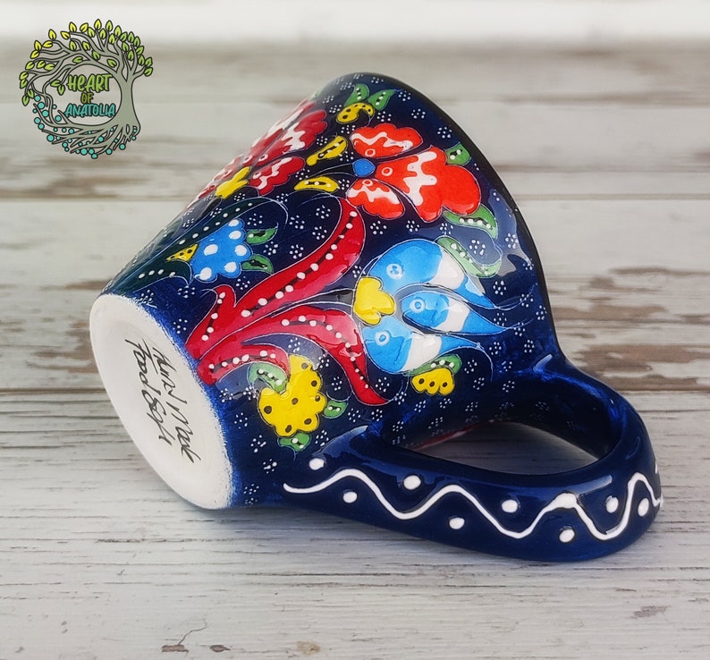 May include: A blue ceramic coffee cup with a floral design in red, yellow, blue, and white. The cup has a handle and the words "Hand Made" are written on the bottom.
